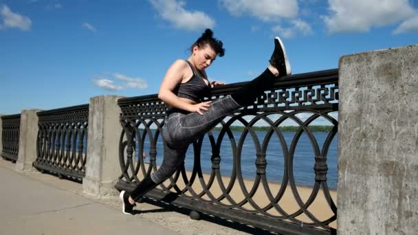 Jeune femme en vêtements de sport étirant les jambes le long de la clôture — Video