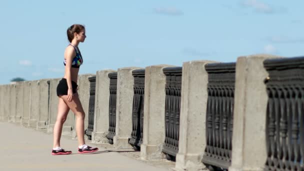 Meisje in sports shirt en broek stretching haar benen na de ochtend joggen — Stockvideo