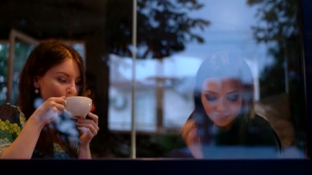 Chicas sentadas junto a la ventana bebiendo café y hablando por teléfono y entre ellas — Vídeos de Stock