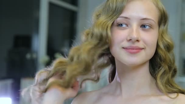 schöne junge Mädchen mit langen lockigen blonden Haaren und blauen Augen sieht sich vor dem Make-up
