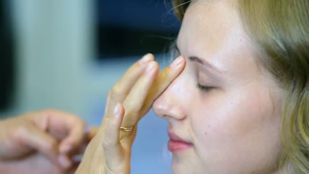 Artista de maquiagem profissional aplicando base de creme para cabelos longos loiros e olhos azuis modelo de pele . — Vídeo de Stock