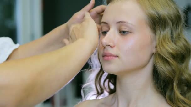 Artista de maquiagem profissional aplicando base de creme para cabelos longos loiros e olhos azuis modelo de pele . — Vídeo de Stock