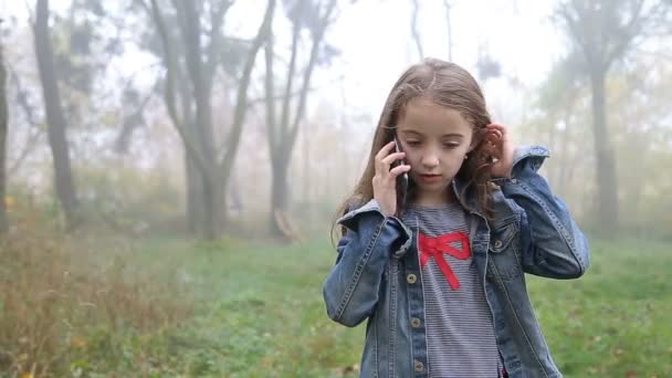 Little European girl with a long hair, blue jacket, black pants, sneakers and blue eyes. A frightened little child is walking through the foggy deserted forest and talking cell phone. Loneliness. — Stock Video