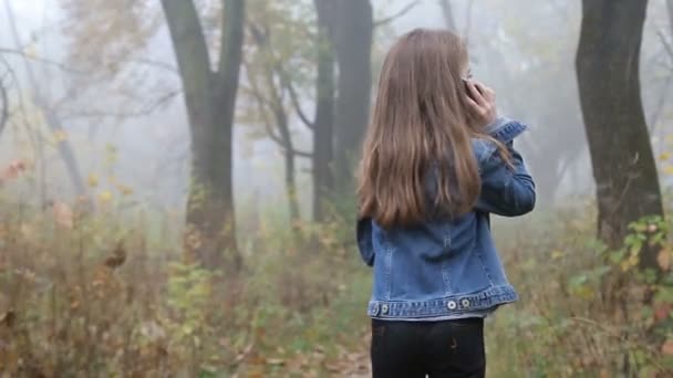 Little European girl with a long hair, blue jacket, black pants, sneakers and blue eyes. A frightened little child is walking through the foggy deserted forest and talking cell phone. Loneliness. — Stock Video