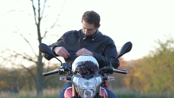 Um jovem de jaqueta de couro preto senta-se na motocicleta, tira óculos de sol e veste o capacete branco antes da viagem ao pôr-do-sol do outono — Vídeo de Stock