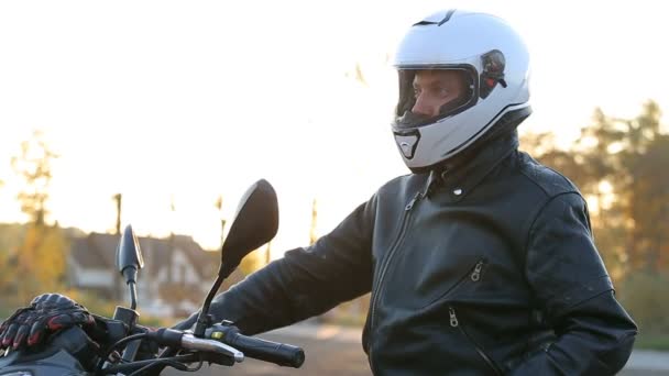 Ein junger Mann in schwarzer Lederjacke sitzt auf einem Motorrad und bereitet seinen weißen Helm vor der Fahrt bei herbstlichem Sonnenuntergang vor — Stockvideo