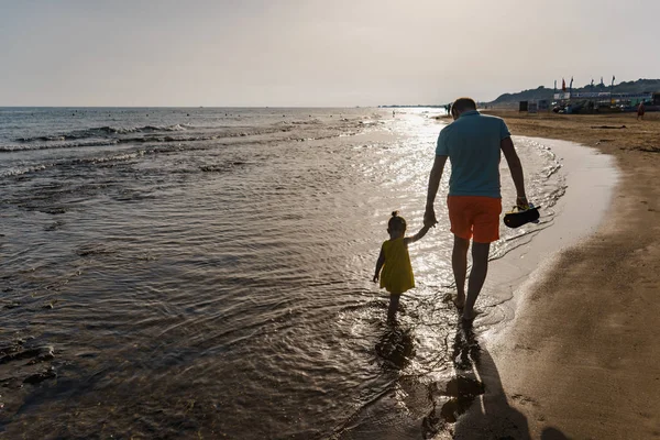 En man med ett barn promenad längs havsstranden vid solnedgången i motljus Royaltyfria Stockbilder