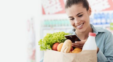 Gülümseyen genç kadın süpermarkette alışveriş, o bir alışveriş çantası elinde ve onun smartphone kullanarak