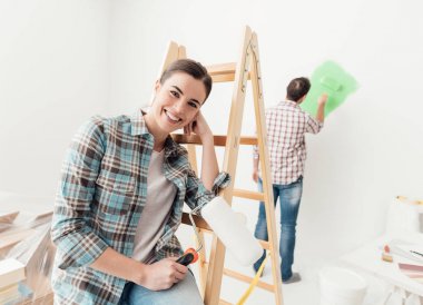 Ev baştan yaratma ve yenileme: Genç çift, onların yeni ev Boyama Boya silindiri adam kullanıyor ve kadın bir merdiven yaslanmış ve gülümseyen