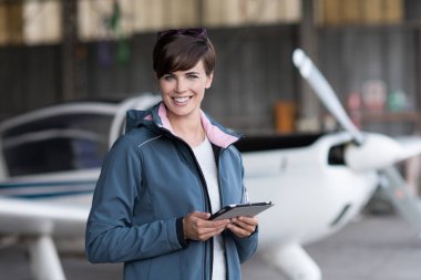 Kendine güvenen kadın pilotu hangar gidiş daha önce hazırlanmasında, o onun tablet ve kamera, arka planda hafif uçak gülümseyen Havacılık apps kullanıyor