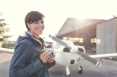 Genç kadın fotoğrafçı ve turist havaalanında gülümseyen, o bir dijital fotoğraf makinesi, pervaneli uçak ve hangar arka planda tutuyor