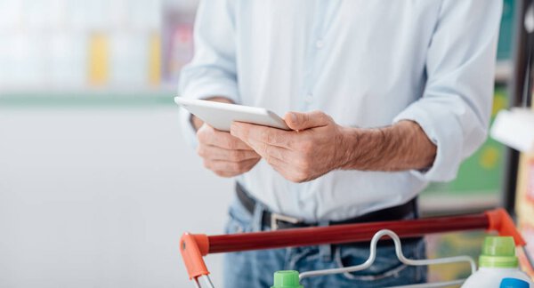 Man doing grocery shopping at the supermarket and using a digital touch screen tablet, he is searching offers and products