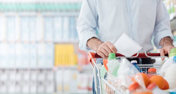 Man at the supermarket shopping with a grocery list and pushing a full cart, lifestyle and retail concept