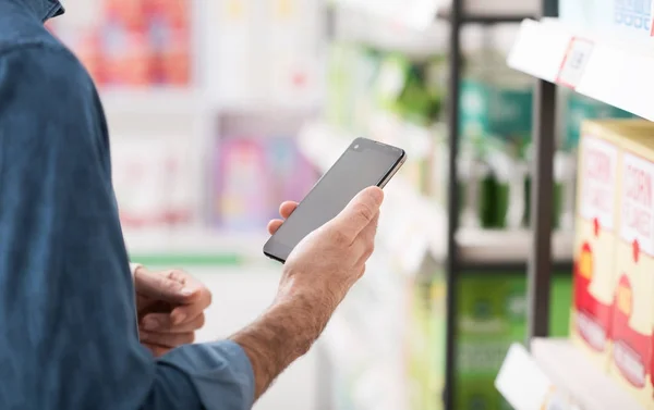 Man shopping at the supermarket, he is searching products on the shelf and using apps on his smartphone, retail and technology concept