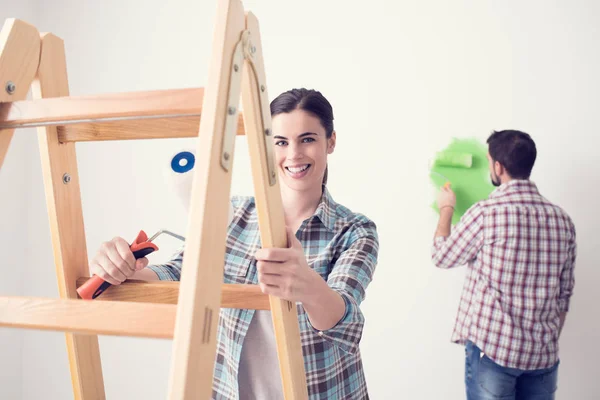 若い女と男の家 絵画の壁を改装 女性がはしごに傾いて カメラに笑顔 — ストック写真