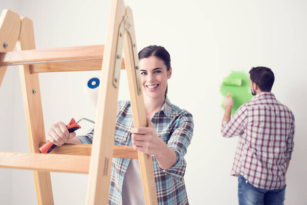Young woman and man renovating their house and painting walls, the woman is leaning on a ladder and smiling at camera