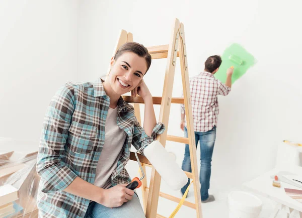 Ev baştan yaratma ve yenileme: Genç çift, onların yeni ev Boyama Boya silindiri adam kullanıyor ve kadın bir merdiven yaslanmış ve gülümseyen
