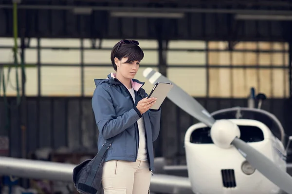 Confident female pilot in the hangar doing a pre-flight preparation, she is connecting and using aviation apps on her digital tablet, propeller plane on the background