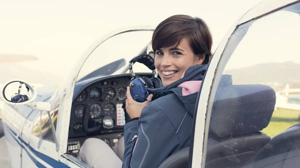 Smiling female pilot in the light aircraft cockpit, she is holding aviator headset and looking at camera