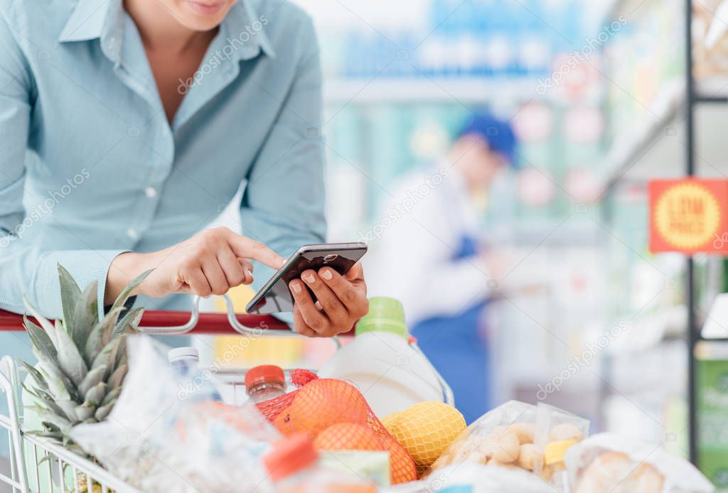 Woman doing grocery shopping at the supermarket, she is leaning on the shopping cart and connecting with her phone, apps and retail concept