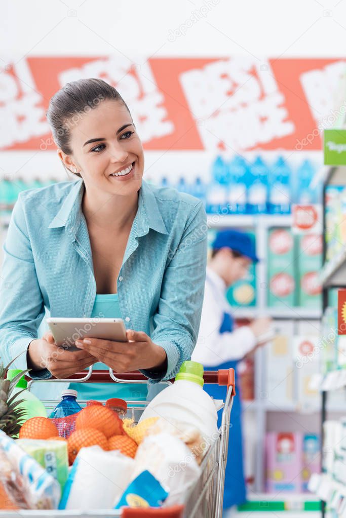 Woman shopping at the supermarket, she is leaning on the shopping cart and using mobile apps on her tablet, technology and retail concept