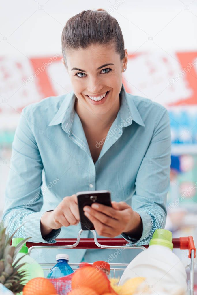 Smiling young woman doing grocery shopping at the supermarket, she is leaning on the shopping cart and connecting with her phone, apps and retail concept