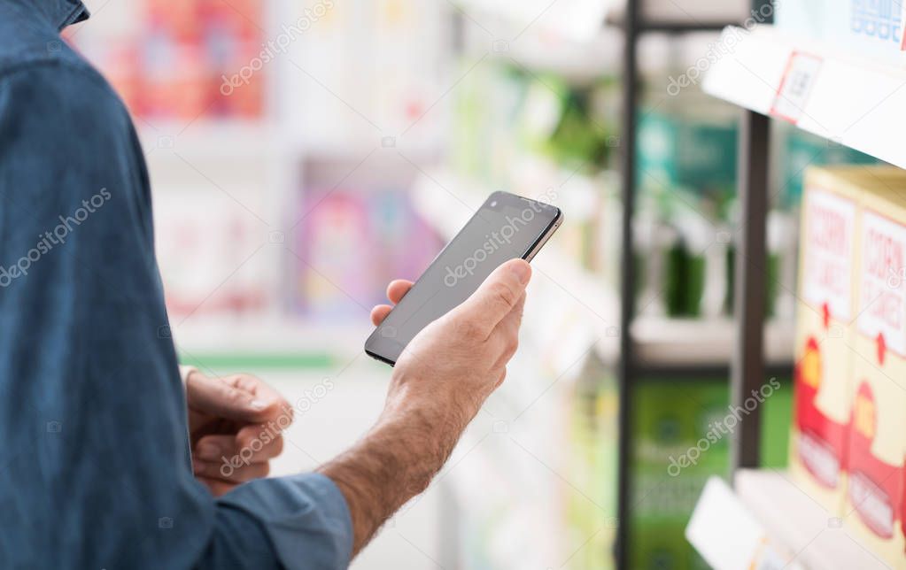 Man shopping at the supermarket, he is searching products on the shelf and using apps on his smartphone, retail and technology concept