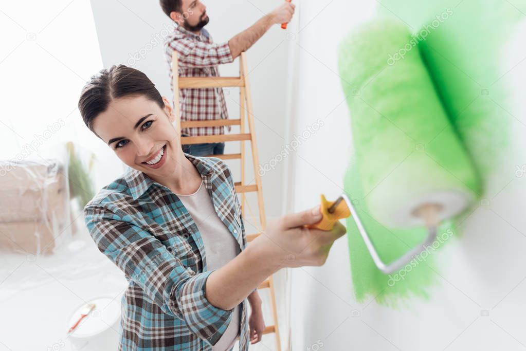 Happy couple painting walls in their new house: the man is standing on a ladder and the woman is using a paint roller and applying bright green paint