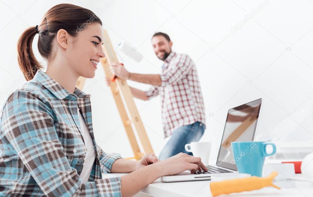 Young smiling couple renovating and remodeling their new apartment, the man is painting the walls with a roller and the woman is connecting with a laptop
