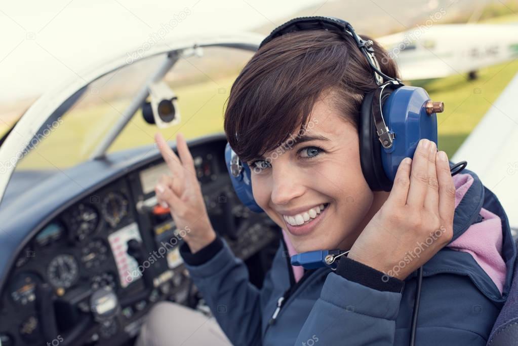 Smiling female pilot in the light aircraft cockpit, she is wearing aviator headset and making a V sign