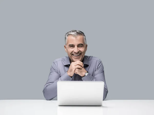 自信的成功微笑的商人坐在办公桌前 用手提电脑工作 他在看摄像头 — 图库照片