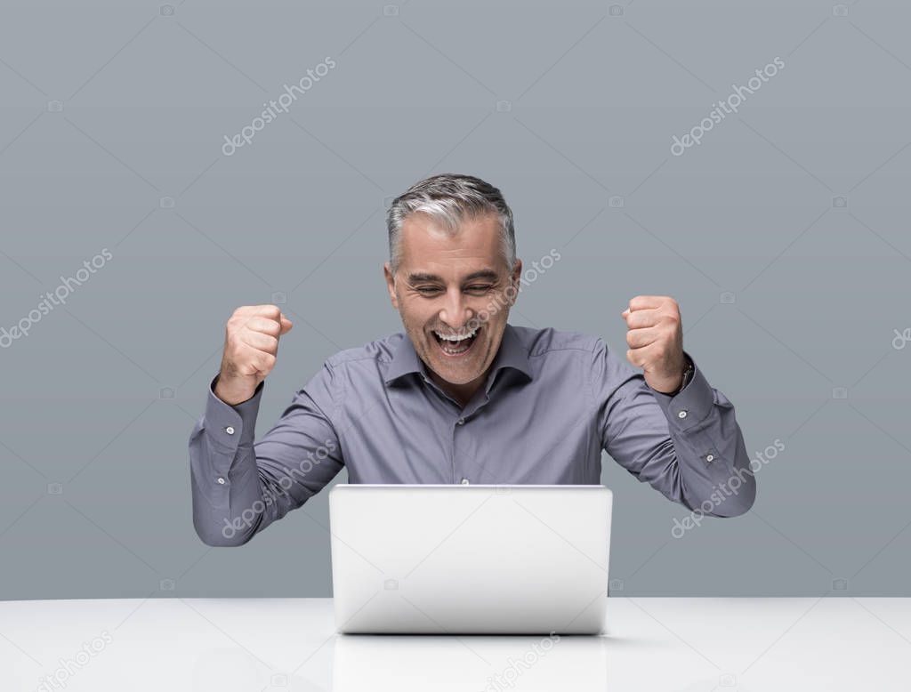 Cheerful mature businessman working at office desk with a laptpop, he is cheering with raised fists, achievement and success concept