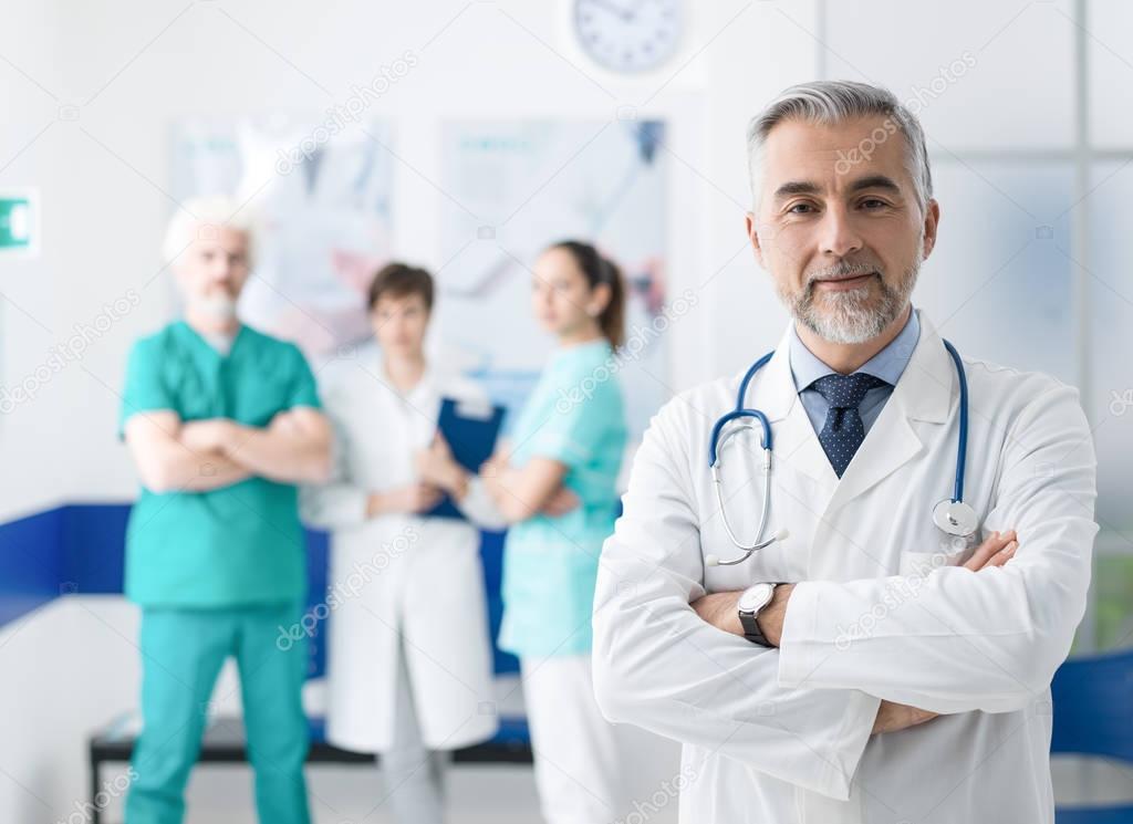 Confident smiling doctor posing and the hospital with arms crossed and medical team working on the background