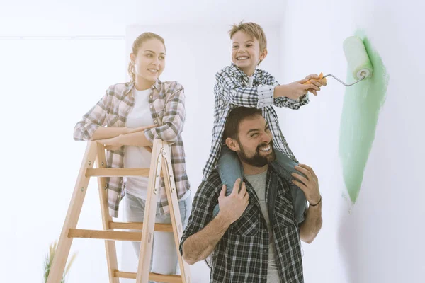 Happy family renovating their new home, the father is piggybacking his son and helping him painting with a roller, the mother is smiling and standing on a ladder
