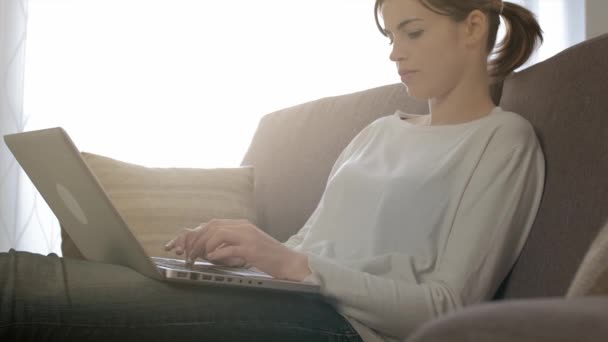 家のソファーに座って若い魅力的な女性 彼女はリラックス ラップトップ上で入力 インターネットへの接続 — ストック動画