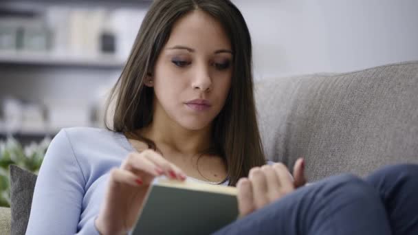 若い魅力的な女性がリラックスできる家で 彼女はソファーに横になっていると 本を読んで 彼女はカメラに微笑みかける — ストック動画