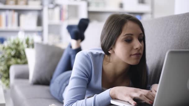 微笑的年轻女孩在家放松在客厅和躺在沙发上 她正在使用笔记本电脑和社交网络 — 图库视频影像