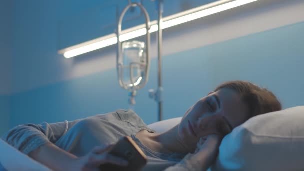 Молодая пациентка лежит в постели в больнице и пользуется смартфоном — стоковое видео