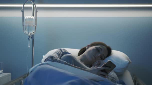 Jonge patiënt ligt in bed in het ziekenhuis en gebruikt haar smartphone — Stockvideo