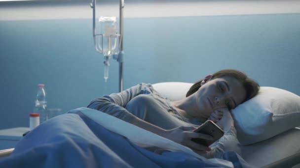Молодая пациентка лежит в постели в больнице и пользуется смартфоном — стоковое видео