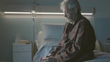 Hastanede yatan yaşlı adam yatakta oturup ilaç alıyor.