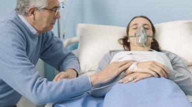 Endişeli bir baba, hasta kızına hastanede yardım ediyor ve elini tutuyor. Yatakta oksijen maskesi takıyor.