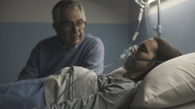 Hastanede kızıyla yan yana oturan ve geceleri ona yardım eden yaşlı adam uyuyor ve oksijen maskesi takıyor.