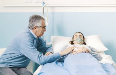 Endişeli bir baba, hasta kızına hastanede yardım ediyor ve elini tutuyor. Yatakta oksijen maskesi takıyor.