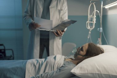 Hastane yatağında oksijen maskesi takmış bir hastaya yardım eden profesyonel doktor tıbbi kayıtları kontrol ediyor.