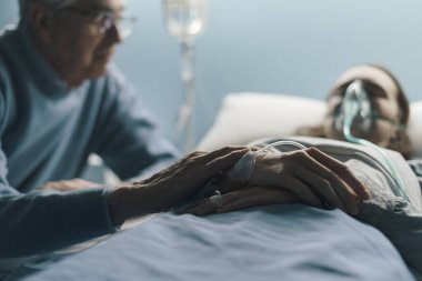 Endişeli kıdemli adam genç kızına hastanede yatağında yardım ediyor, elini tutuyor ve onun yanında oturuyor.