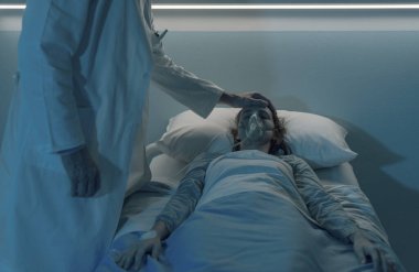 Doktor, gece hastanede yatan bir hastaya yardım ederken ve alnına dokunurken oksijen maskesiyle yatakta yatıyor.