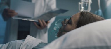 Hastane yatağında oksijen maskesi takmış bir hastaya yardım eden profesyonel doktor tıbbi kayıtları kontrol ediyor.