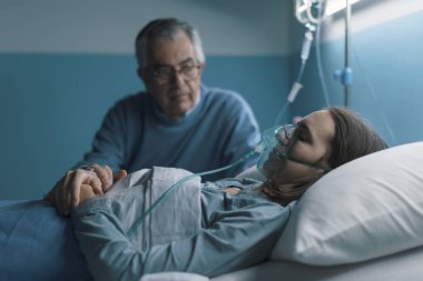 Endişeli kıdemli adam genç kızına hastanede yatağında yardım ediyor, elini tutuyor ve onun yanında oturuyor.