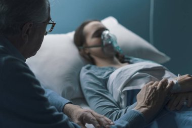 Endişeli kıdemli adam genç kızına hastanede yatağında yardım ediyor, elini tutuyor ve onun yanında oturuyor.